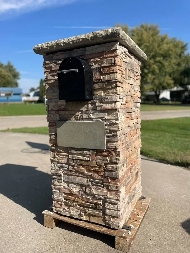 Stacked-stone mailbox column with a rough stone cap, a black mailbox and a blank rectangular plaque, standing on a wooden pallet in a driveway.