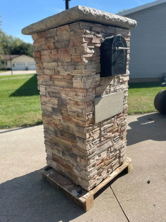 Stacked-stone mailbox column, seen from the side, with a rough stone cap, a black mailbox and a blank rectangular plaque, on a wooden pallet.
