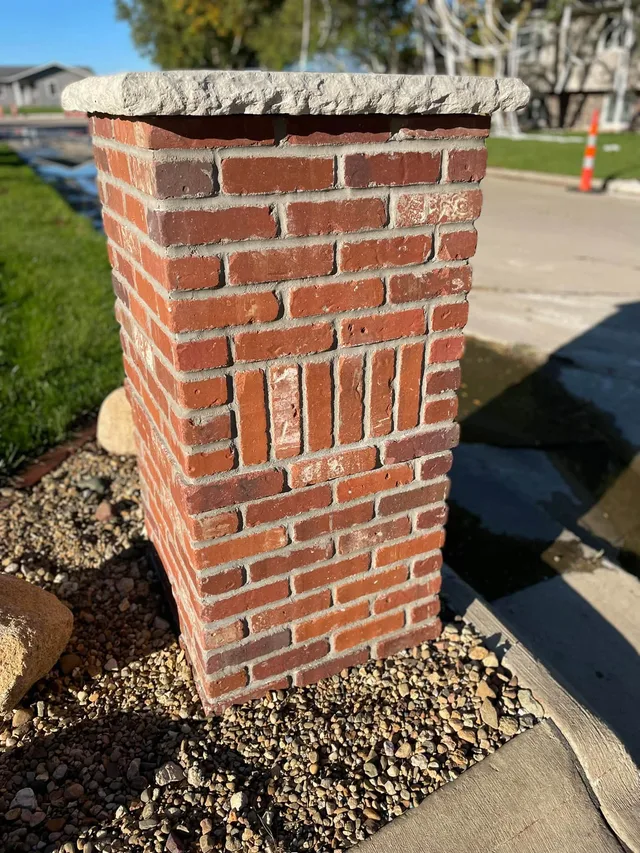Red brick column with a rough stone cap and a vertical-brick panel, set in a gravel bed beside a curb.
