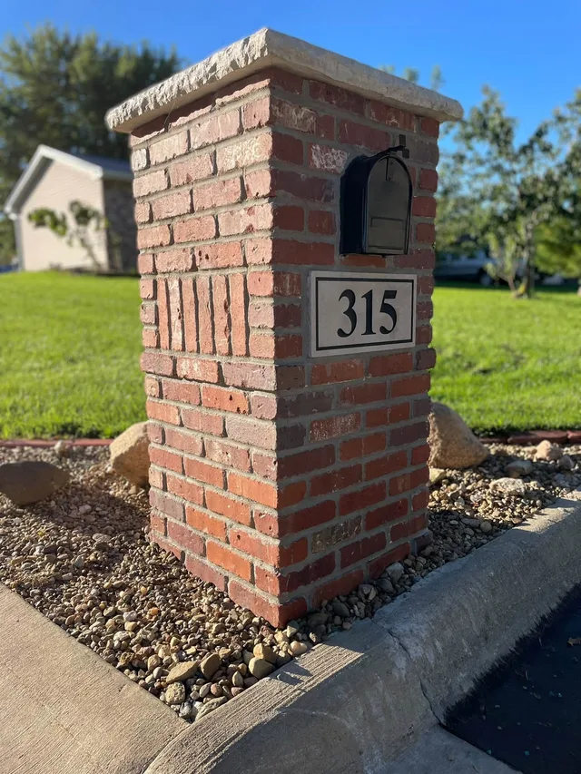 Red brick mailbox column with a rough stone cap, a black mailbox, and a framed plaque reading 315, set in a gravel bed beside a curb.